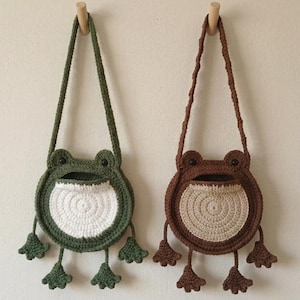 Könnte beinhalten: Zwei gehäkelte Umhängetaschen in Froschform. Eine ist grün mit weißem Bauch, die andere braun mit beigem Bauch. Beide haben lange Träger und Froschfüße. Sie hängen an Holzdübeln an einer weißen Wand.