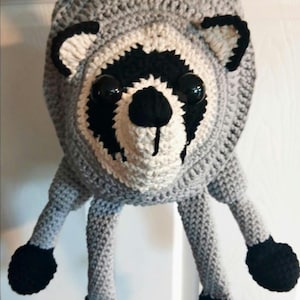 May include: A crochet bag shaped like a raccoon head with a grey body, white face markings, black eye patches, a black nose, and two ears. The bag features four dangling grey legs with black paws and a strap for carrying, crafted from yarn.