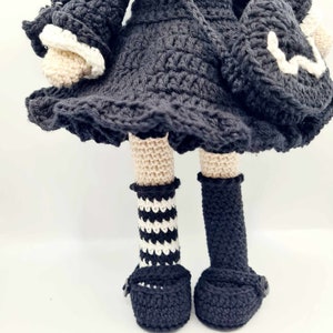 May include: A crocheted doll wearing a black dress with white trim, black and white striped socks, and black shoes.