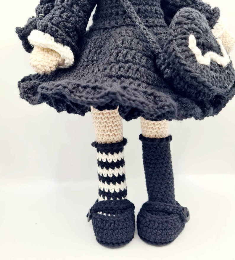May include: A crocheted doll wearing a black dress with white trim, black and white striped socks, and black shoes.