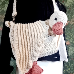 May include: Crocheted goose-shaped shoulder bag in cream and pink. The bag features a white goose head with a pink beak and feet. The body of the bag is cream-colored with a ribbed texture and a matching strap. The bag is attached to a silver clasp.