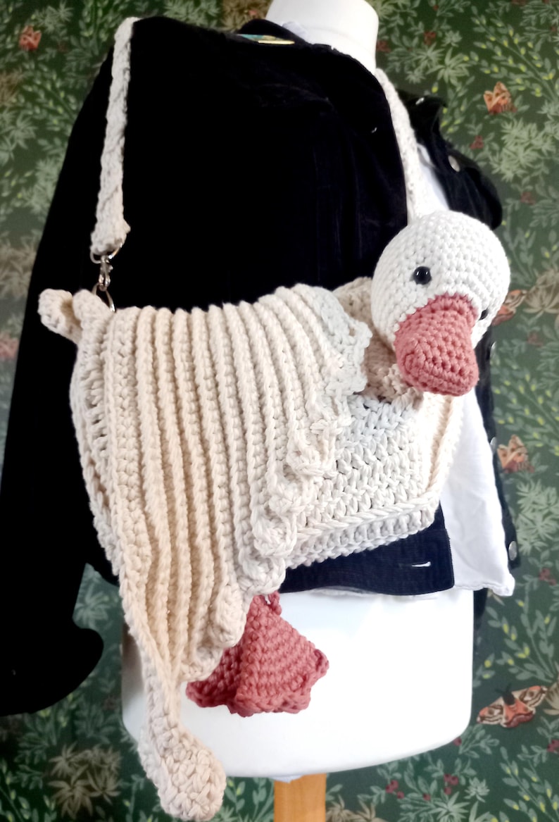 May include: Crocheted goose-shaped shoulder bag in cream and pink. The bag features a white goose head with a pink beak and feet. The body of the bag is cream-colored with a ribbed texture and a matching strap. The bag is attached to a silver clasp.