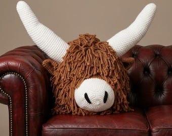 May include: A handmade crochet pillow shaped like a Highland cow's head. The pillow features a shaggy brown mane, a white face, and large white horns. It is resting on a dark red leather couch.