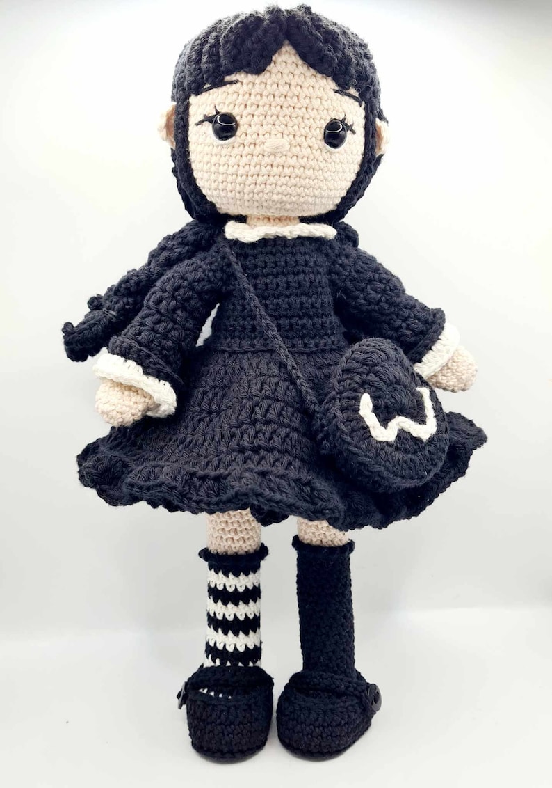 May include: A crocheted doll with black hair, a black dress, white socks with black stripes, and black shoes. The doll is holding a black crocheted bag with a white letter W on it.