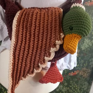 May include: A handmade crochet duck-shaped bag in shades of brown, green, and orange. The bag features a detailed duck head with an orange beak and a green head. The wings are brown with a cream-colored trim. The bag has a shoulder strap.