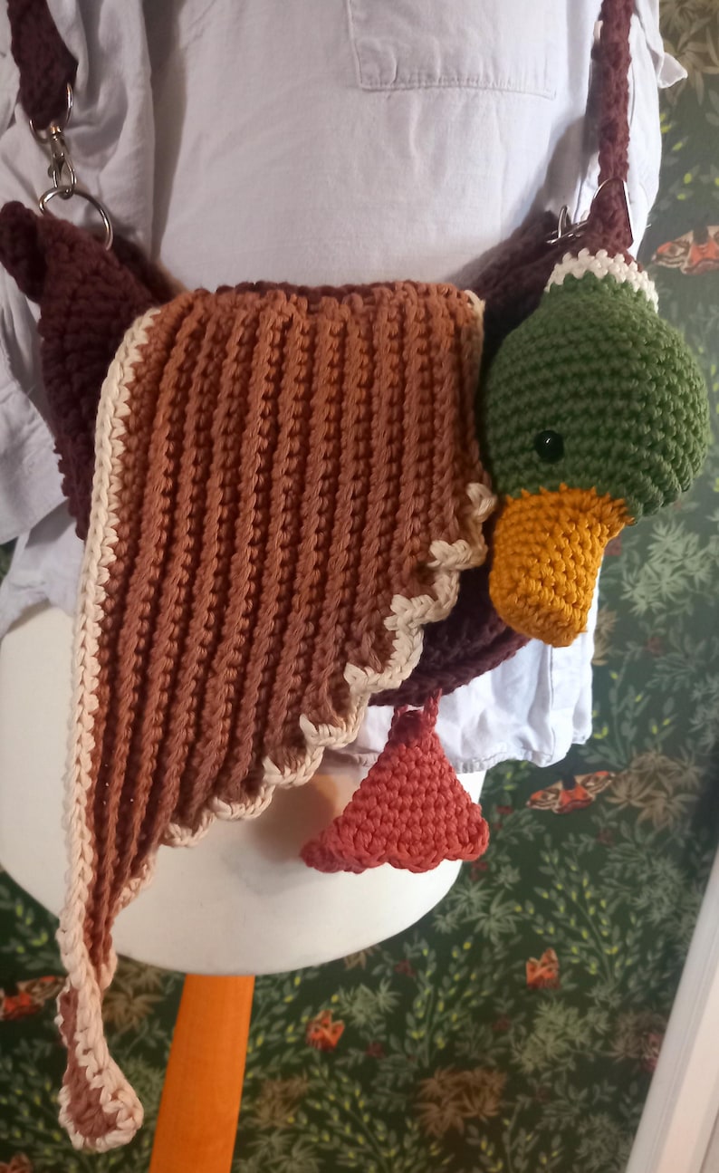May include: A handmade crochet duck-shaped bag in shades of brown, green, and orange. The bag features a detailed duck head with an orange beak and a green head. The wings are brown with a cream-colored trim. The bag has a shoulder strap.