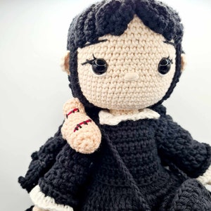 May include: A crocheted doll with black hair, a white collar, and a black dress. The doll is holding a crocheted arm in its right hand.