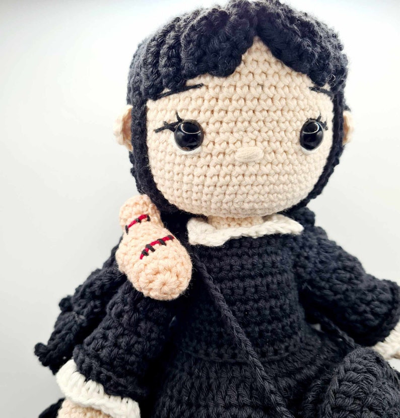 May include: A crocheted doll with black hair, a white collar, and a black dress. The doll is holding a crocheted arm in its right hand.