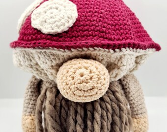 May include: A handmade crochet gnome doll with a burgundy mushroom cap adorned with white spots. The gnome has a beige face and nose, with a long, braided brown beard. The arms and feet are beige.