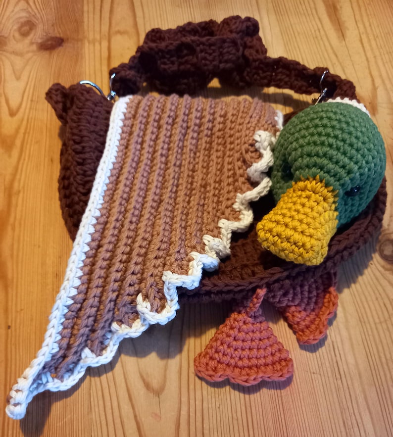 May include: A handmade crochet bag shaped like a duck. The bag features a brown body, a tan and white patterned flap, and a green duck head with a yellow beak. Orange feet are attached to the bottom. The bag has a brown handle.