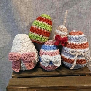 May include: A collection of five crocheted Easter egg decorations. The eggs are in various colors, including white, green, red, blue, and pink, and are adorned with bows and decorative twine. They are displayed on a wooden crate.