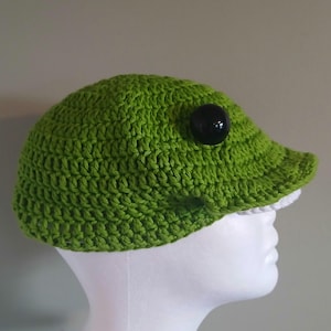 May include: A bright green crocheted cap featuring a prominent, large, round, glossy black button on the side. The hat has a short, stiffened brim and a textured, yarn-based construction, displayed on a plain white mannequin head form.