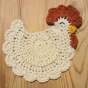 May include: A crochet coaster shaped like a chicken, featuring a cream-colored body with a scalloped edge. The head has a rust-colored comb and wattle, a yellow beak, and a single black embroidered eye. The design is flat and circular in the center.
