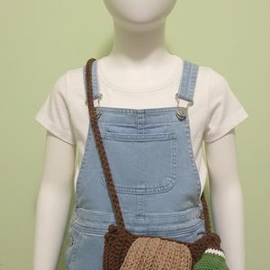 May include: A brown crochet duck-shaped crossbody bag with a long strap. The bag features a tan wing, a green head with a white accent, and a yellow foot. The bag is worn by a mannequin wearing denim overalls and a white t-shirt.