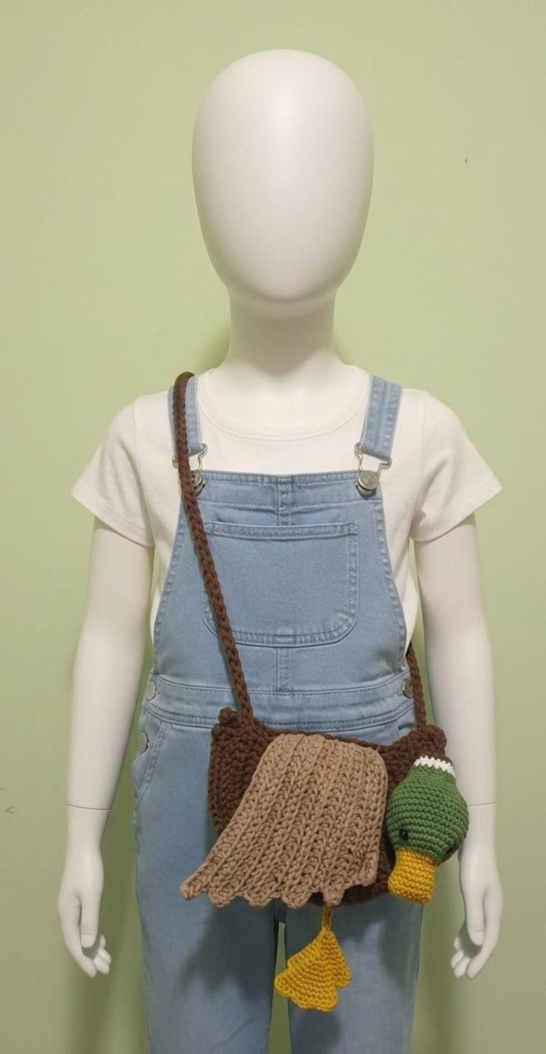 May include: A brown crochet duck-shaped crossbody bag with a long strap. The bag features a tan wing, a green head with a white accent, and a yellow foot. The bag is worn by a mannequin wearing denim overalls and a white t-shirt.