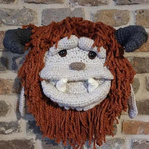 May include: A handmade crochet wall hanging featuring a whimsical monster face. The face is primarily light gray with brown eyes, a tan nose, and two white tusks. The monster has shaggy, rust-colored hair and gray horns.