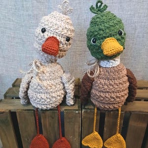 Op de afbeelding: Twee handgemaakte gehaakte eendenspeeltjes. Eén is beige met een oranje snavel en poten, de andere is bruin en groen met een gele snavel en poten. Beide hebben zwarte ogen en zitten op een houten kist. Een leuke toevoeging voor de kinderkamer.