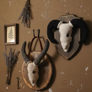 May include: Crocheted ram skull wall decor with dark grey horns and a cream-coloured skull, mounted on a wooden plaque. The skull is displayed on a brown wall with other decorative items, including dried lavender and a framed fern print.