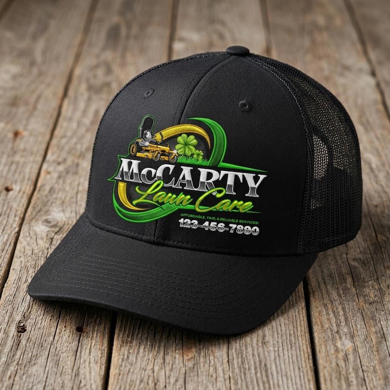Black trucker cap with embroidered lawn care logo showing yellow zero turn mower, green shamrock and gold green ribbon design on rustic wood surface — landscaper hat branding for grass cutting and yard service businesses