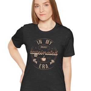May include: A dark heather grey t-shirt with a brown graphic that says "In My Highly Caffeinated Era" with a coffee cup icon.