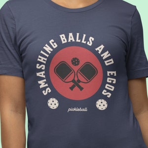 May include: A navy blue t-shirt with a white circle graphic that says "Smashing Balls and Egos" with a red circle inside that has two crossed pickleball paddles and a pickleball. The text "pickleball" is written in white below the red circle.