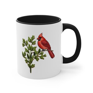 May include: A white ceramic mug with a black rim and handle. The mug features a red cardinal perched on a green branch.