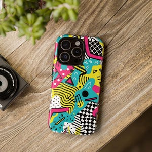 May include: A colorful phone case with a vibrant abstract design. The case features a mix of shapes, including triangles, lines, and checkered patterns, in bright colors like pink, yellow, and teal. The phone case is on a wooden surface.