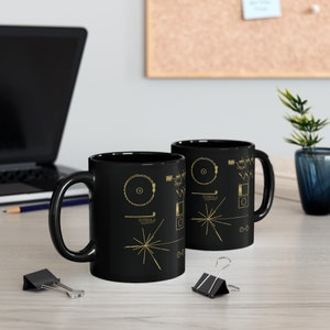 May include: Two black ceramic mugs with gold-colored Voyager Golden Record designs. The designs include a diagram of the solar system, a pulsar map, and other scientific illustrations. The mugs are on a light-colored wooden surface.