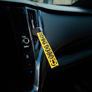 May include: A yellow keychain tag with the text "PULL TO EJECT" in black, hanging in a car. The tag is rectangular with black arrows. The car's interior is black with carbon fiber accents.