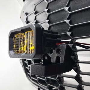 May include: A black and yellow LED light mounted on a black bracket. The light is attached to a black grille with a honeycomb pattern.