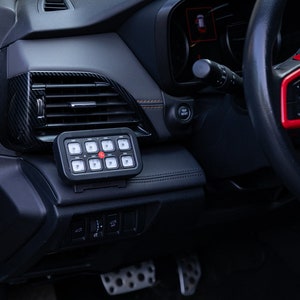 May include: A black rectangular eight-button switch panel mounted on the dashboard of a vehicle. The panel features eight square buttons with white icons and a central red button. The dashboard includes carbon fiber trim and a steering wheel.
