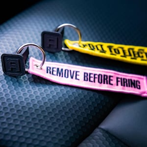 May include: Three keychains with black square tags featuring the letter "A" and metal rings. One keychain has a pink tag with "REMOVE BEFORE FIRING" in white, and another has a yellow tag with black text.