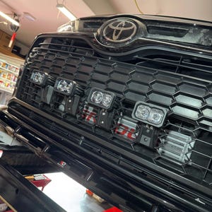 May include: A black Toyota vehicle front grille featuring a honeycomb pattern and four rectangular LED auxiliary lights mounted horizontally. The Toyota emblem is centered at the top, and a metallic intercooler is visible behind the grille mesh.