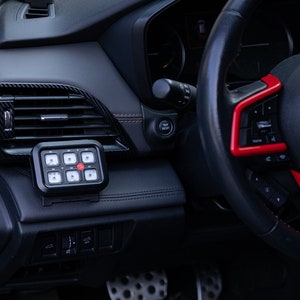May include: A black car interior with a black and red steering wheel. A black button panel with a red button is mounted on the dashboard.