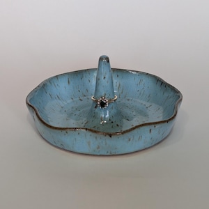 May include: A light blue ceramic ring dish with a cone-shaped center for holding rings. The dish has a scalloped edge and a speckled glaze.