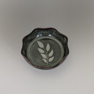 May include: A small, round, ceramic bowl with a wavy rim. The bowl is dark green with a lighter green interior. A white leaf design is in the center of the bowl.