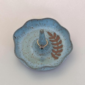 May include: A ceramic ring holder with a light blue speckled glaze and a brown leaf design. The holder has a raised center for rings and a scalloped edge. A gold ring with a black gem is on the holder.