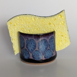 May include: A handmade ceramic sponge holder with a blue leaf pattern and a yellow sponge. The holder has a dark purple rim and a black base. The sponge is inserted into the holder.