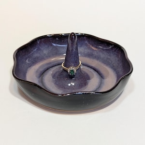 May include: A handcrafted ceramic ring holder with a dark blue and purple glaze. The dish has a wavy rim and a central cone for holding rings. A gold ring with a green gemstone is displayed on the cone.