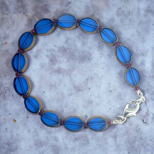 Puede incluir: Un brazalete de vidrio azul con cierre dorado y detalles rojos. El brazalete está hecho de cuentas ovaladas.