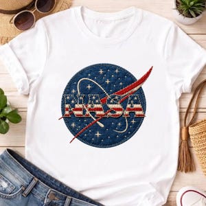 May include: A white short-sleeved t-shirt featuring a circular graphic on the chest. The graphic displays the text NASA in a bold font, filled with a red and white striped pattern, set against a dark blue background with white stars.