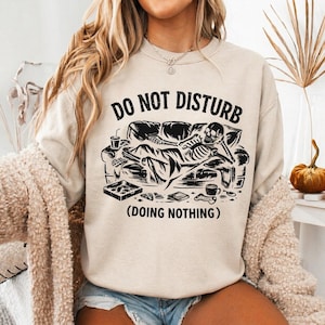 Puede incluir: Sudadera beige con un gráfico negro de un esqueleto descansando en un sofá, con las palabras "DO NOT DISTURB" arriba y "(DOING NOTHING)" abajo. El esqueleto está rodeado de objetos como una bebida y un libro.