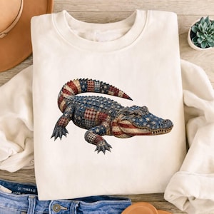 May include: A cream-colored crewneck sweatshirt featuring a graphic of an alligator with a textured pattern resembling the American flag, including red and white stripes and blue sections with white stars across its body and tail.