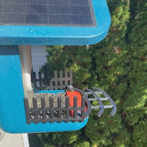 May include: A blue bird feeder with a solar panel on top. The feeder has a black fence-like structure around the seed compartment and a silver perch. The feeder is filled with seeds and has an orange component.