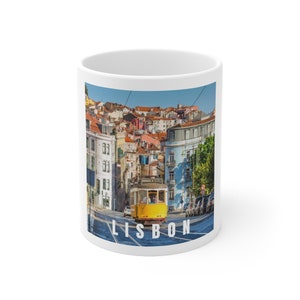 May include: White ceramic mug featuring a vibrant image of Lisbon, Portugal. The design showcases a yellow tram traveling down a street lined with colorful buildings. The word "LISBON" is printed in white at the bottom of the mug.