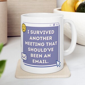 Puede incluir: Taza de cerámica blanca con un diseño morado y azul. La taza tiene un texto que dice "Sobreviví a otra reunión que debería haber sido un correo electrónico."