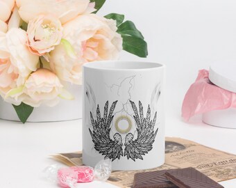 May include: White ceramic mug with a black and white illustration of a pair of wings with a sun in the center. The wings are detailed with feathers and the sun is surrounded by a circle of light.