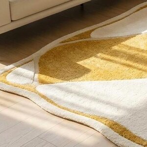 Pu&ograve; includere: Un tappeto morbido e astratto con un design a forma di farfalla. Il tappeto presenta una combinazione di tonalit&agrave; bianche e gialle, con un bordo giallo. Il tappeto &egrave; posizionato su un pavimento in legno chiaro, con una porzione di un divano beige visibile sullo sfondo.
