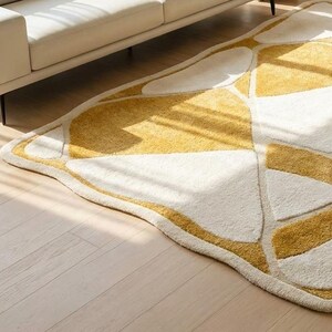 Pu&ograve; includere: Un tappeto a forma di foglia dal design moderno. Presenta un motivo di forme geometriche bianche e gialle, bordate di giallo. Il tappeto &egrave; posizionato su un pavimento in legno chiaro, con un divano bianco sullo sfondo.