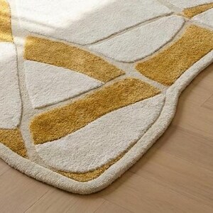 Pu&ograve; includere: Primo piano di un moderno tappeto con un design geometrico. Il tappeto presenta uno sfondo color crema con sezioni giallo senape. I bordi sono arrotondati e il tappeto &egrave; posato su un pavimento in legno chiaro.